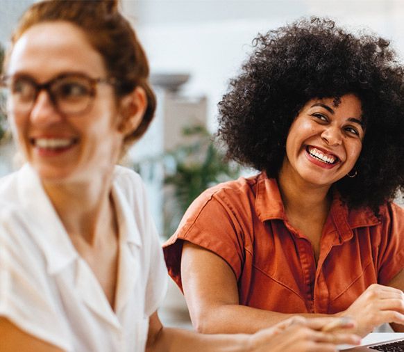 Duas mulheres estão sorrindo enquanto estão sentadas à mesa com um laptop. Quero Mais Performance