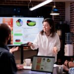 mulher branca de pé à frente de uma tela com gráficos conversando com pessoas sentadas