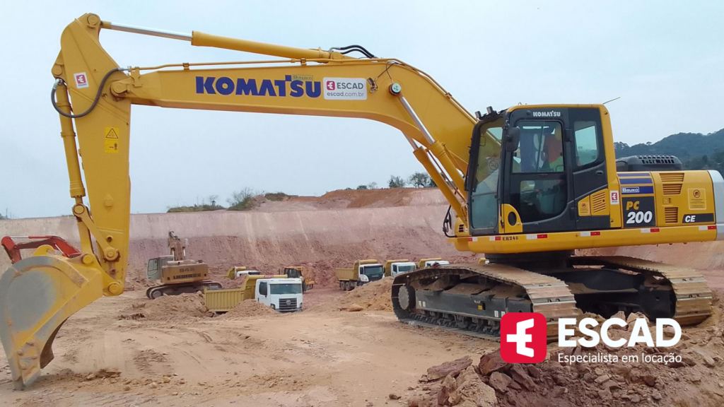 máquina escavadeira de grande porte em primeiro plano e caminhões grandes ao fundo.