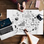 mesa de madeira com notebook e um grande papel com desenhos relacionados ao marketing